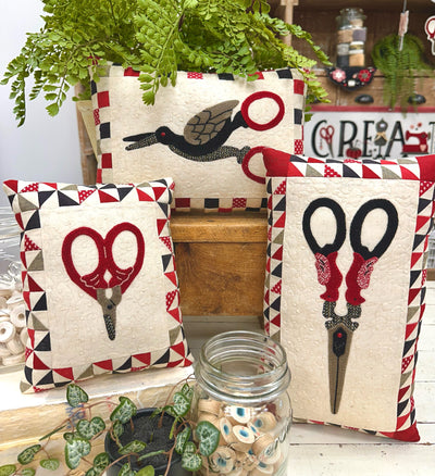 Decorative cotton pillows with woolen vintage scissors design on a wooden surface with plants and jars.