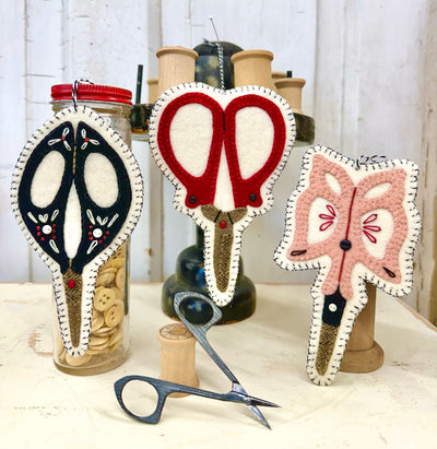 Decorative woolen vintage scissor ornaments on wooden spools with a jar of pins in the background.