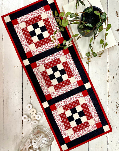 Quilted table runner with red, black, and white pattern on a wooden surface.